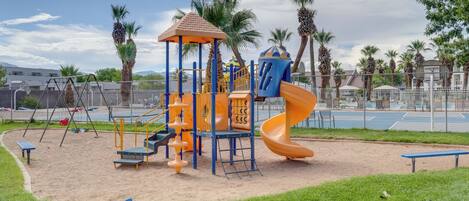 A playground with a slide and climbing structures, surrounded by palm trees and a fence.