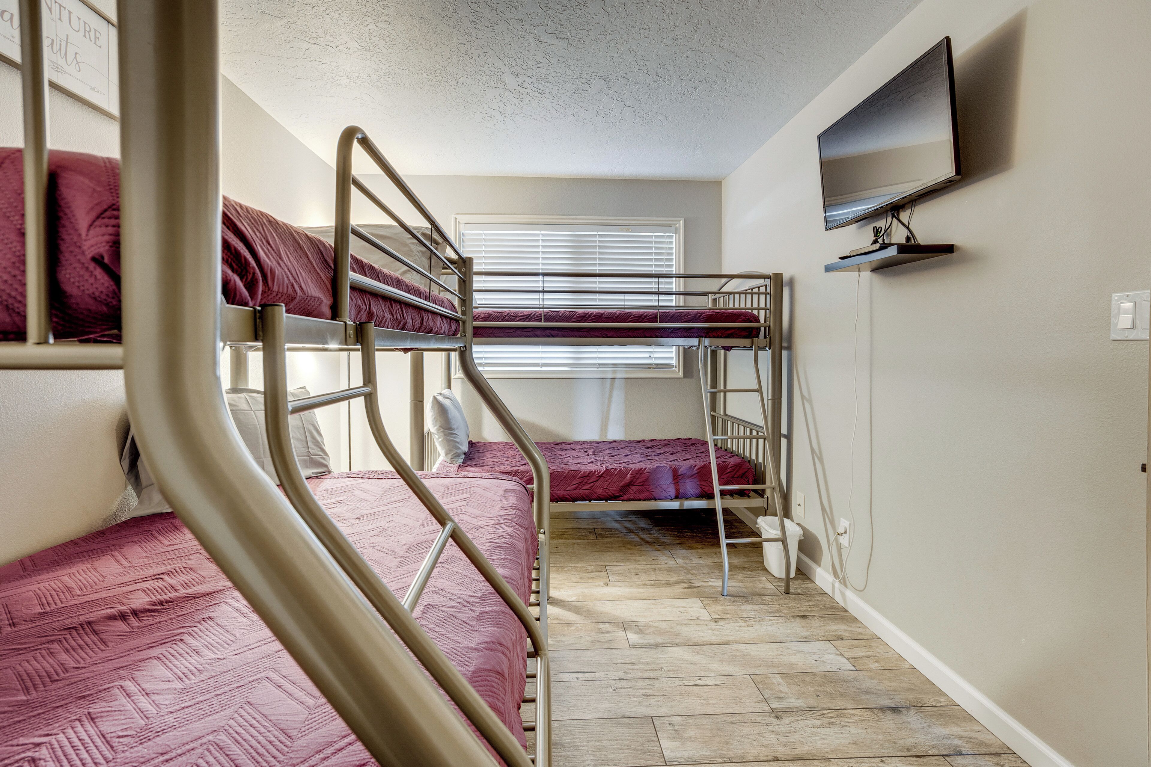 A dormitory room with bunk beds, a television mounted on the wall, and a window with blinds.