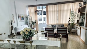 A modern living room with a glass dining table, wooden chairs, a sofa, and a kitchen area with plants. - Spacious 3-bedroom apartment in Copacabana, 3 blocks from the beach. (Rio de Janeiro)