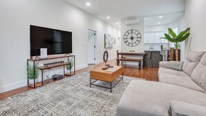 Una sala de estar moderna con una televisión de pantalla plana, una mesa de centro y un sofá. Se ve una cocina al fondo, con gabinetes blancos, y un comedor con una mesa y sillas.