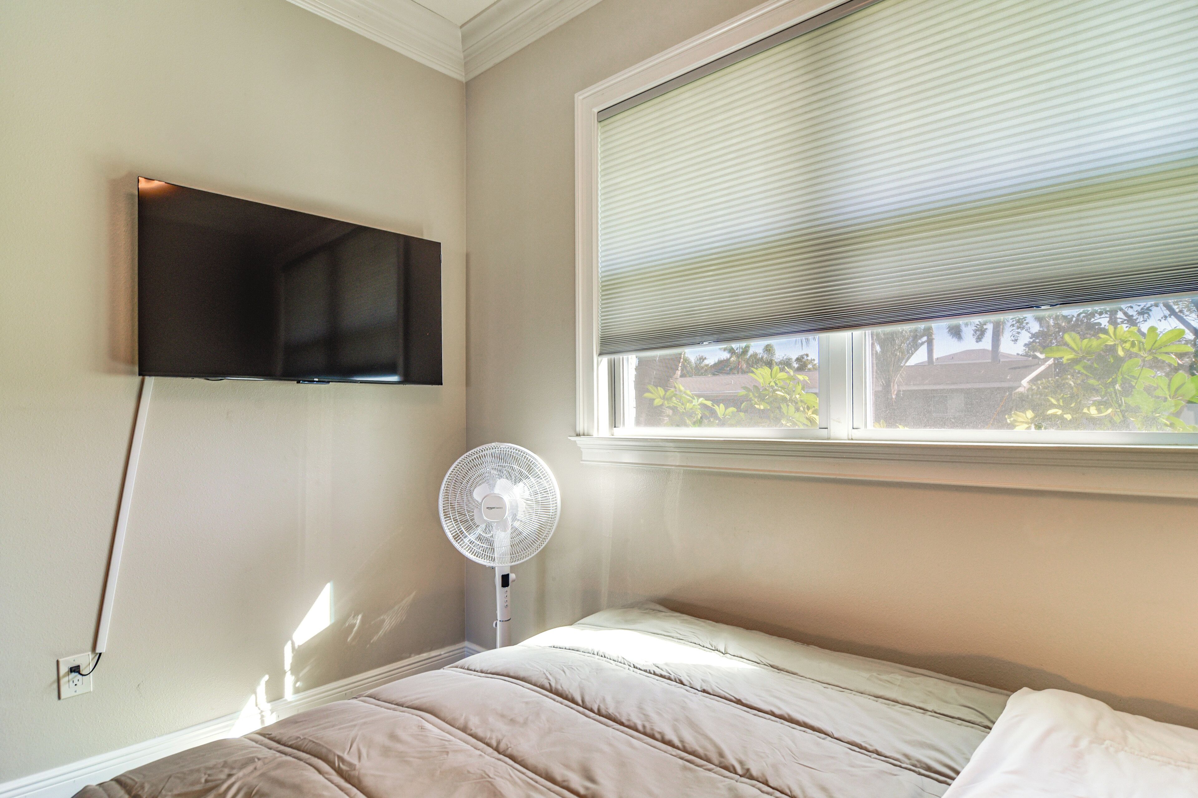A bedroom with a bed, a mounted TV, a standing fan, and a window with blinds.