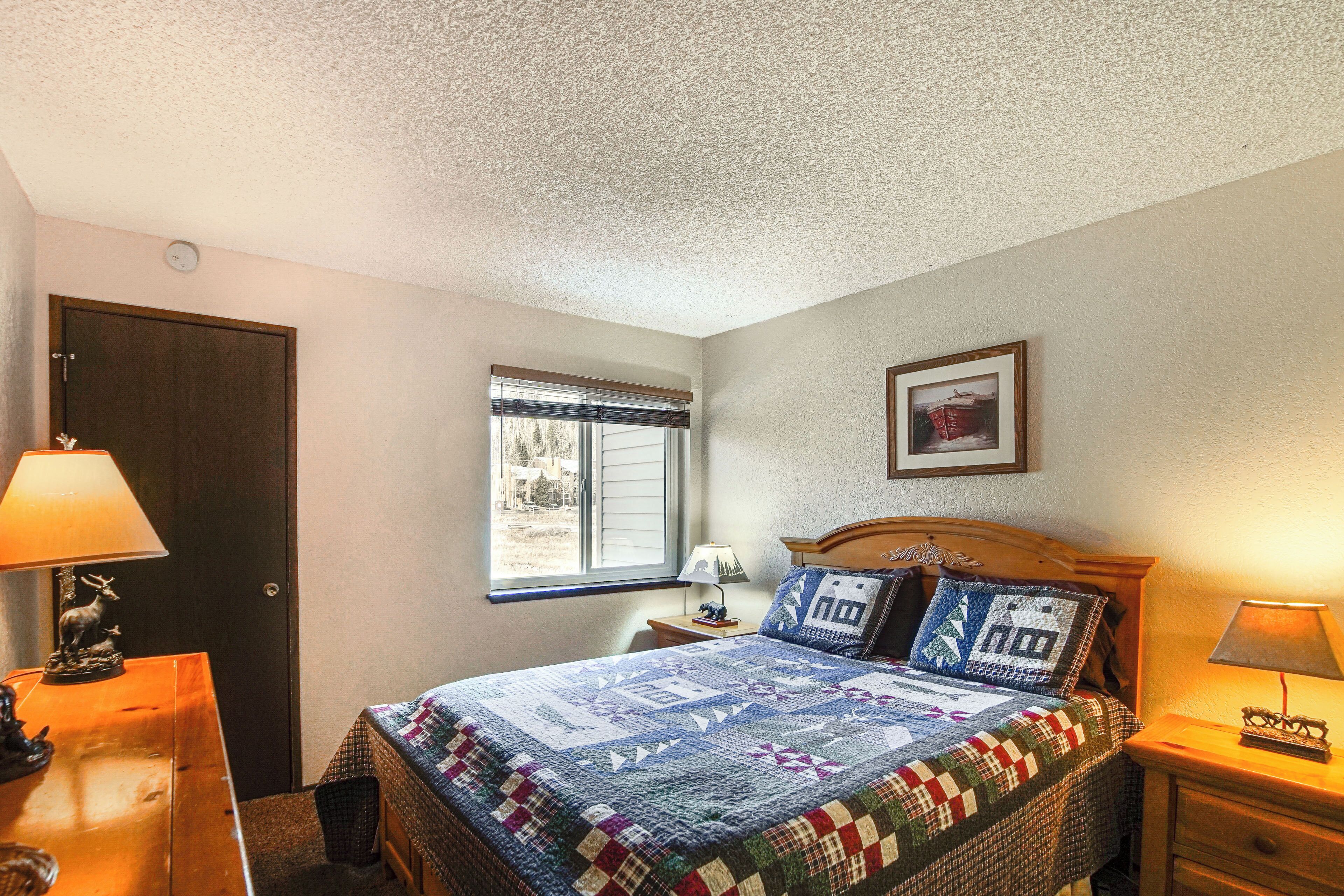 A bedroom with a bed, a window, a door, and a nightstand with a lamp.