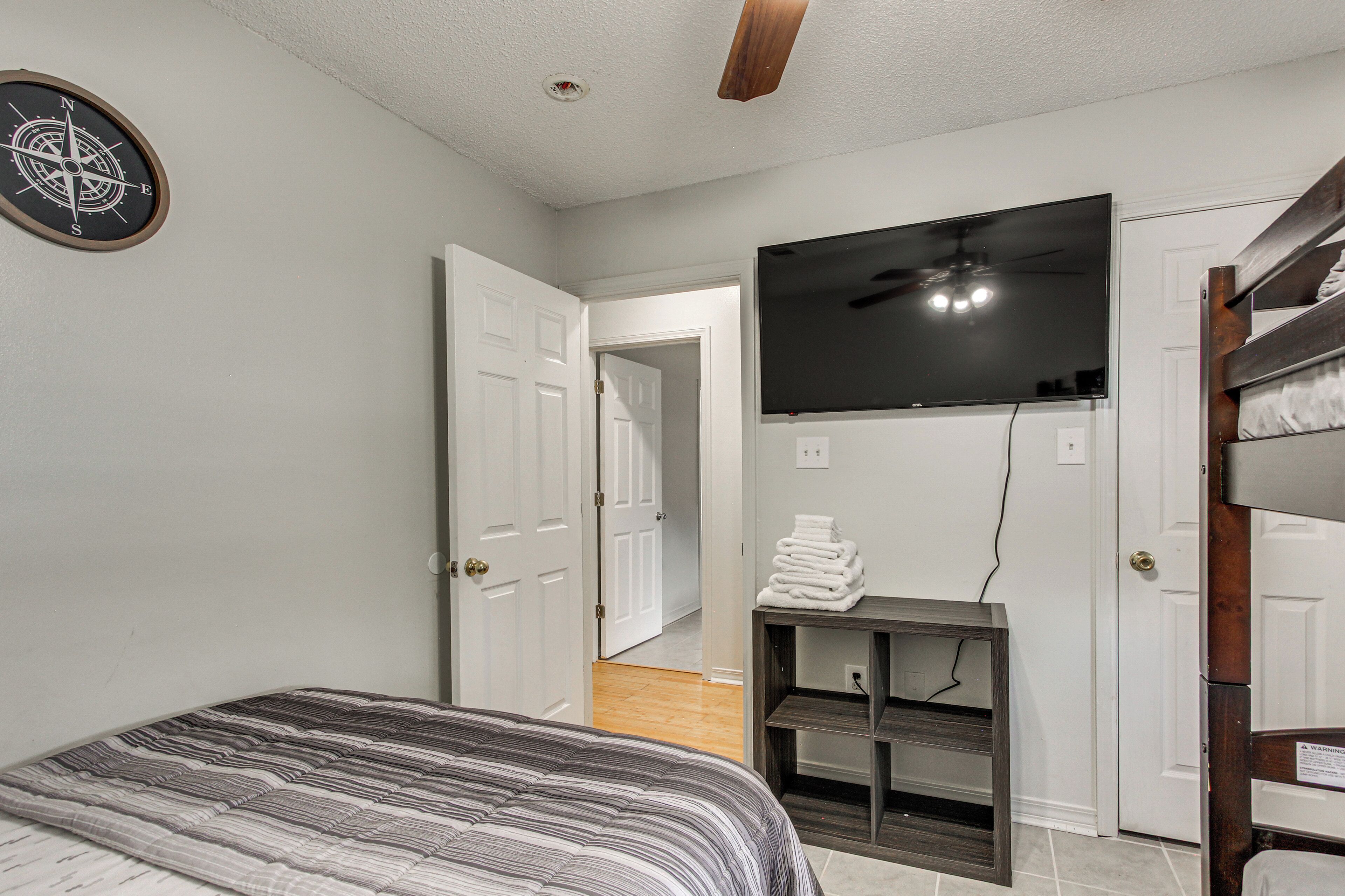 A bedroom with a bed, a TV mounted on the wall, a ceiling fan, and a door leading to another room.