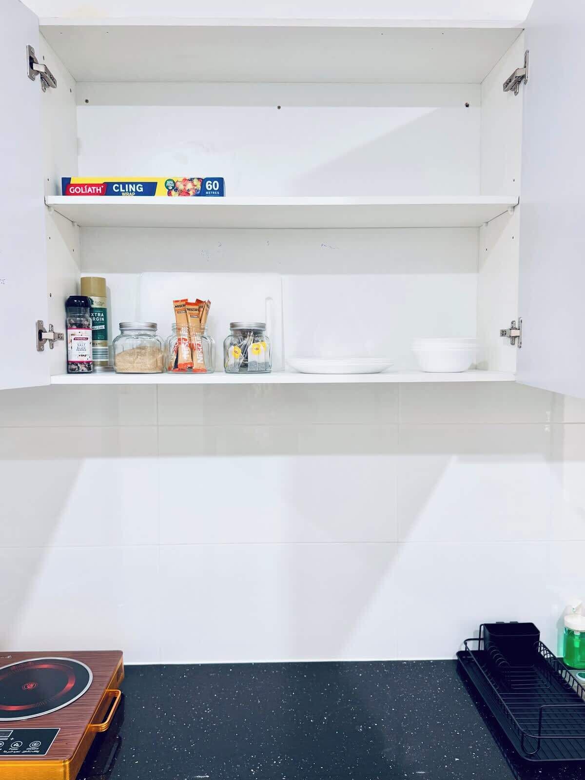 A kitchen with white cabinets, a countertop, and a black backsplash. There is a box labeled 'GOLIATH CLING WRAP' and a '60 METRES' sign on the upper shelf.
