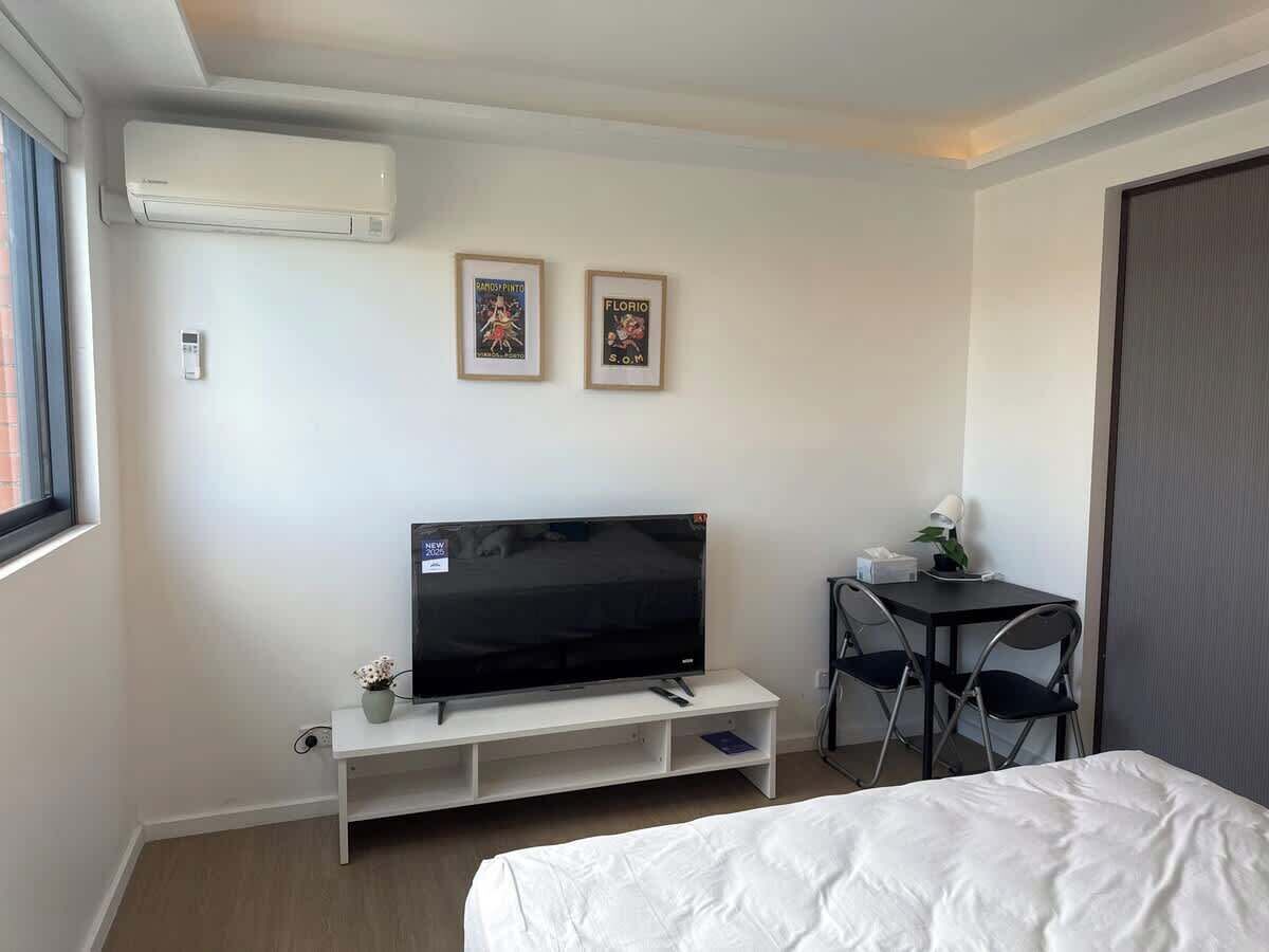 A hotel room with a bed, a TV on a white stand, a desk with a chair, and two framed pictures on the wall.