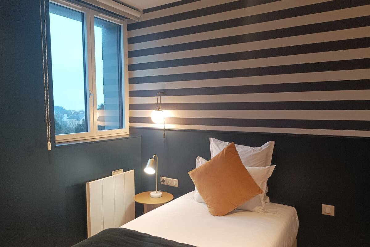 Un dormitorio con cama, mesita de noche, lámpara y ventana.