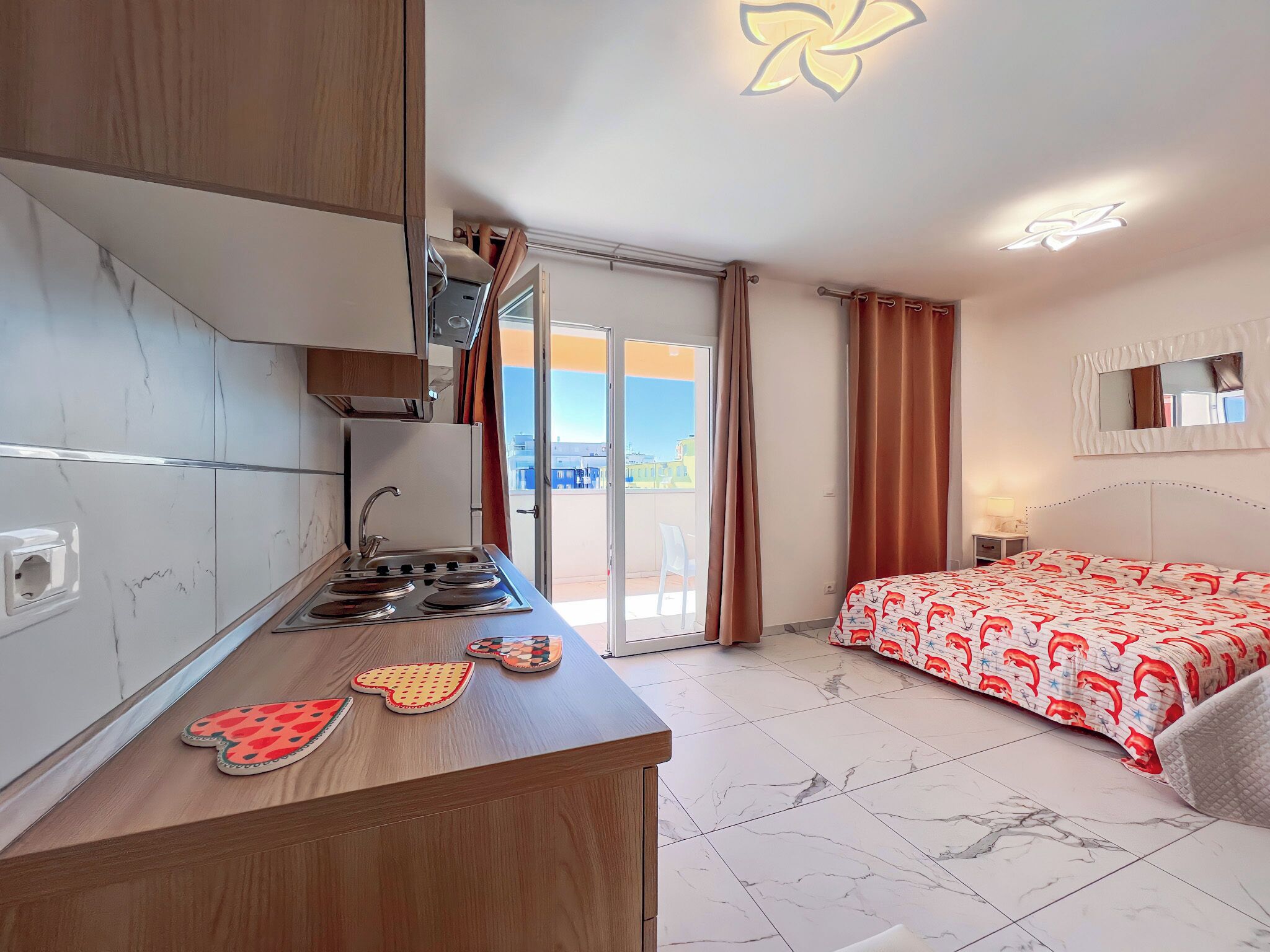 Een kleine hotelkamer met kitchenette, een bed met decoratieve spreien, een grote spiegel en uitzicht door de glazen deur.