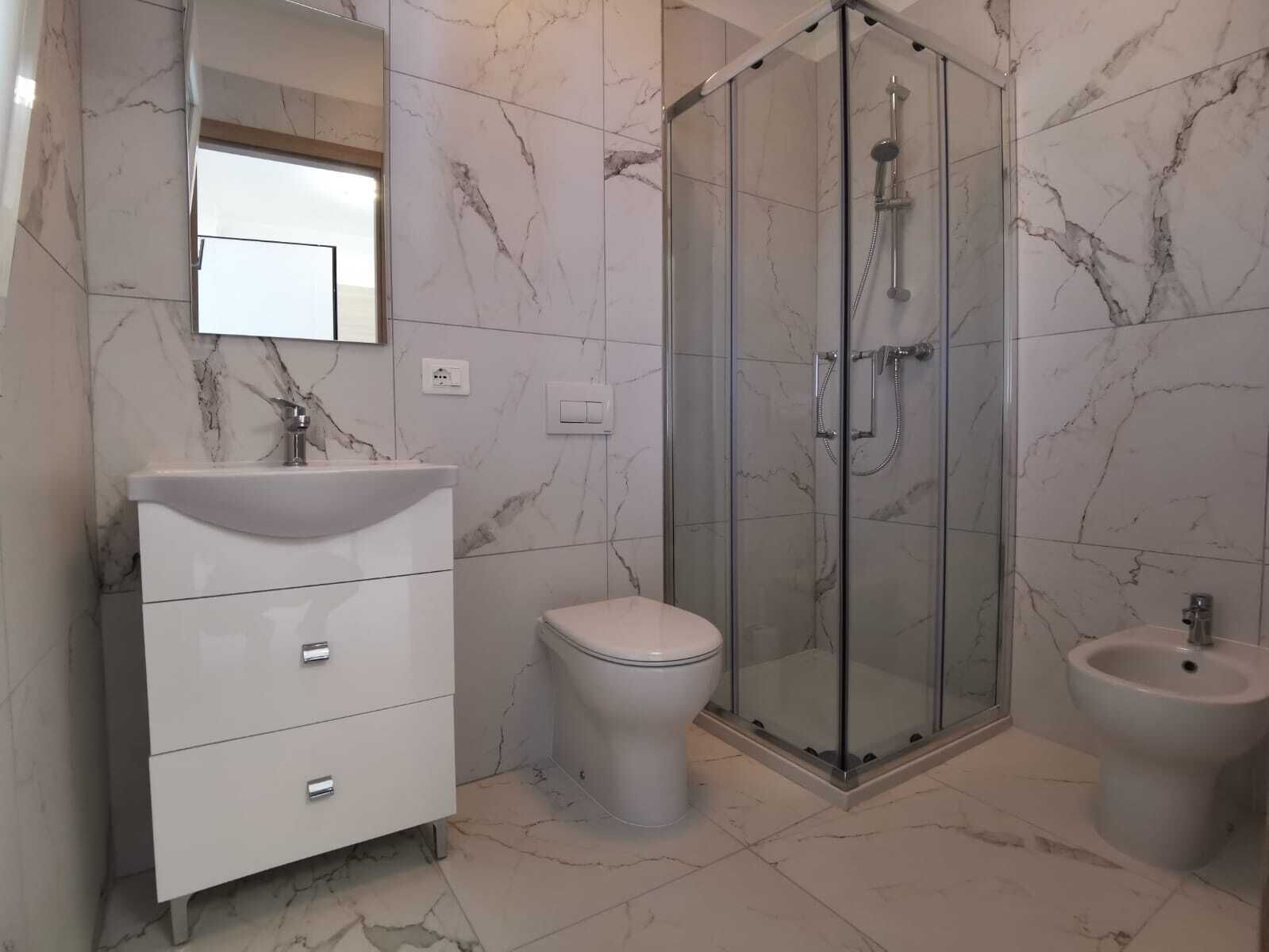 A modern bathroom with a marble floor, a white vanity with drawers, a toilet, a glass-enclosed shower, and a bidet.