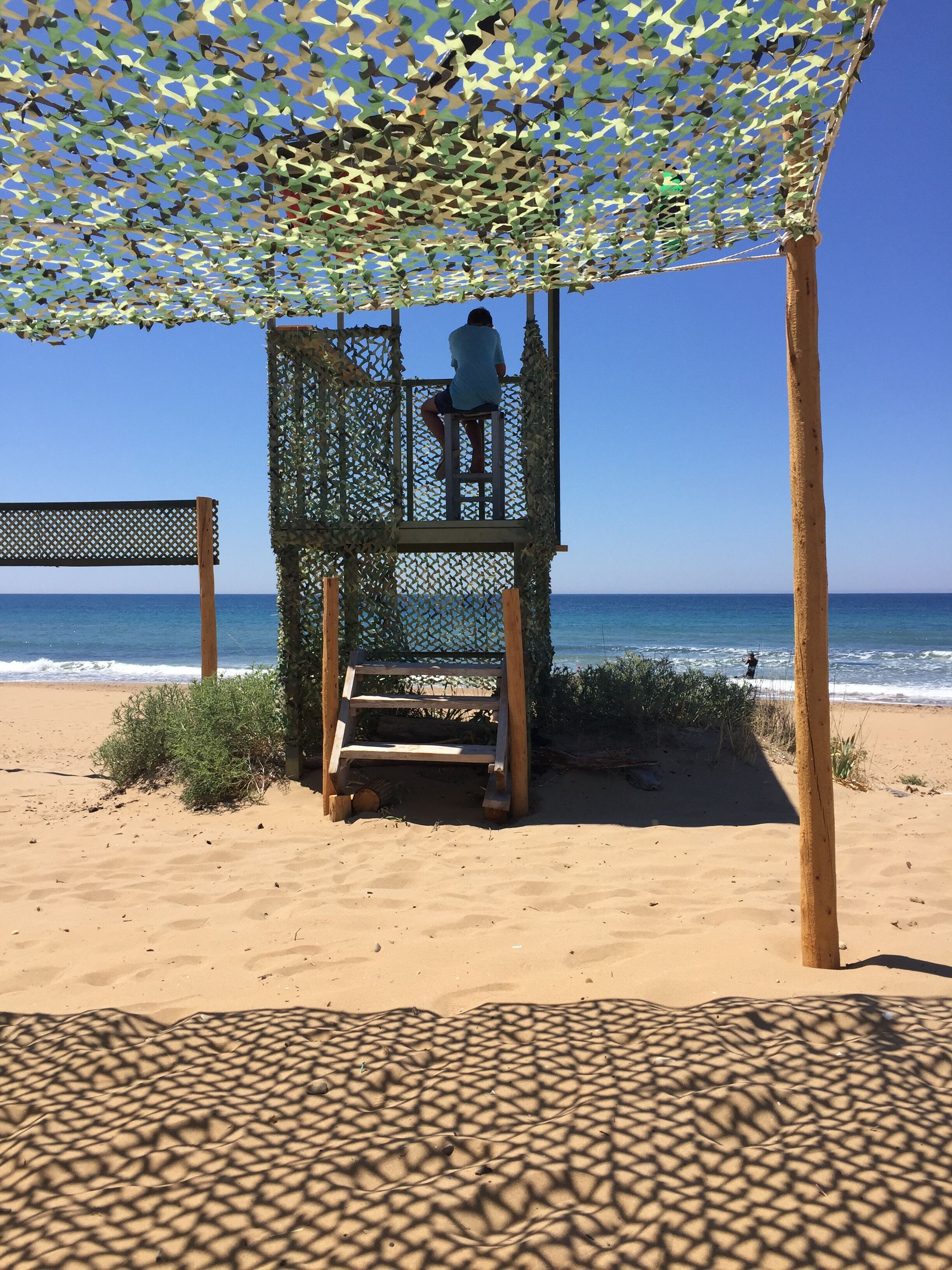 A wooden lifeguard tower with a metal mesh canopy on a sandy beach.