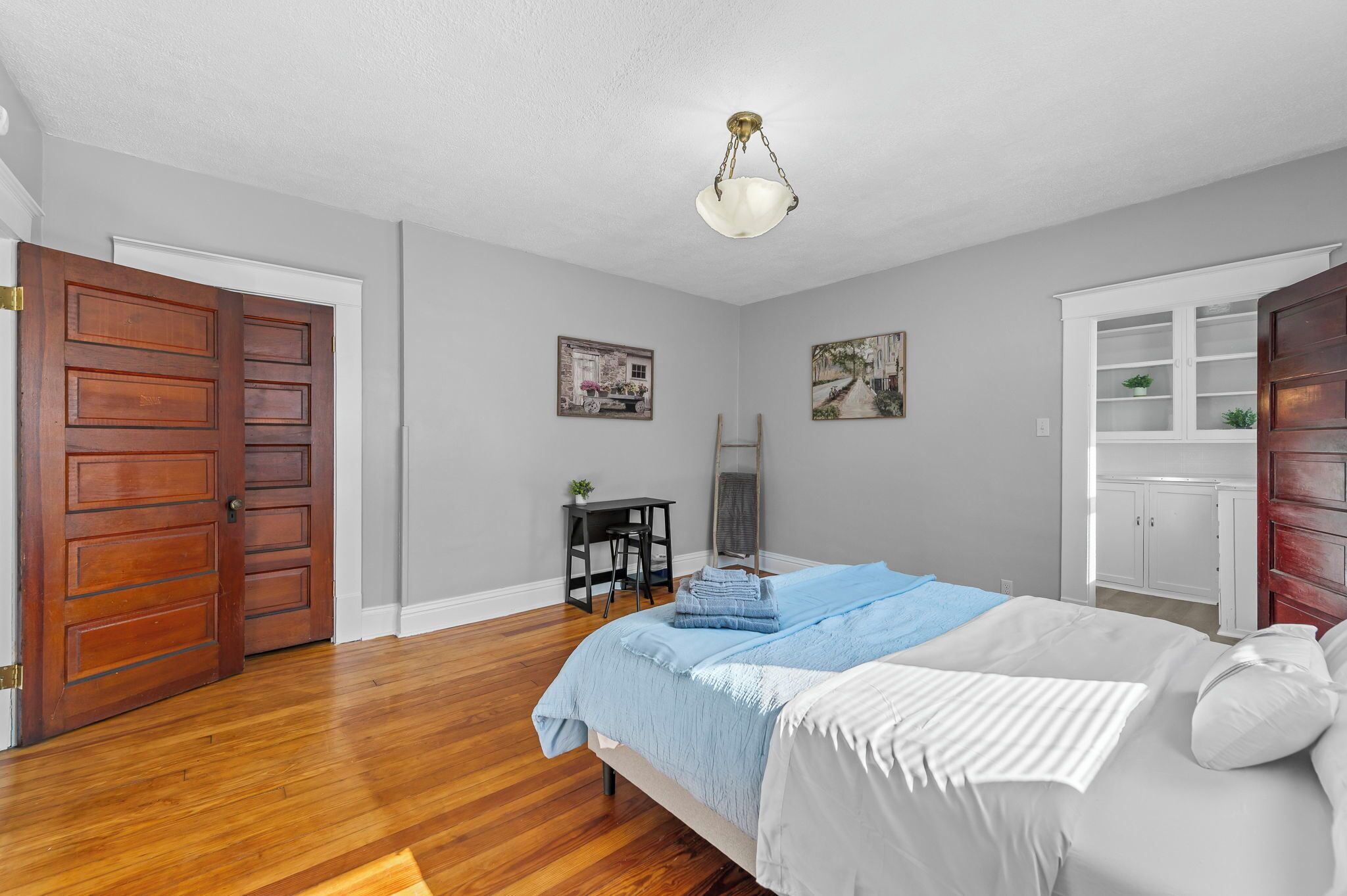 A bedroom with a wooden floor, a bed with white and blue bedding, a desk with a chair, and a wardrobe.