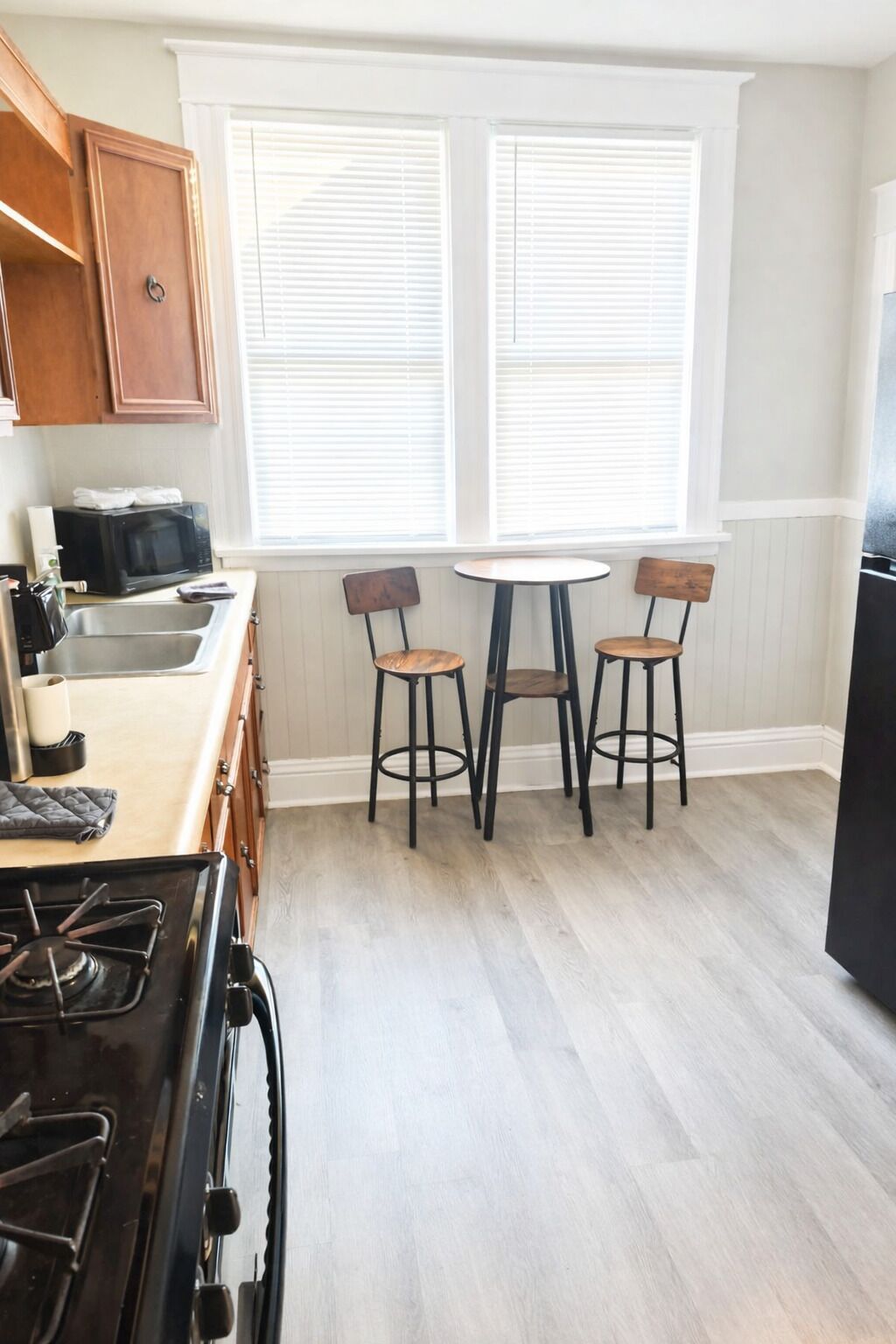 A kitchen with a stove, sink, and two bar stools. There are two windows with blinds, and a refrigerator is partially visible.