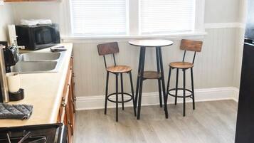 A kitchen with a stove, sink, and two bar stools. There are two windows with blinds, and a refrigerator is partially visible.