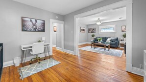 A modern living room with hardwood floors, a gray sofa, and a coffee table. There is a white desk with a chair and a rug in the foreground. - The Maple Attic – Bright 3BR Retreat Near Downtown (Saint Joseph)