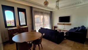 A modern living room with a round wooden table, two sofas, a TV mounted on the wall, and large windows with curtains. - Comfortable poolside apartment near the medina (Marrakesh)