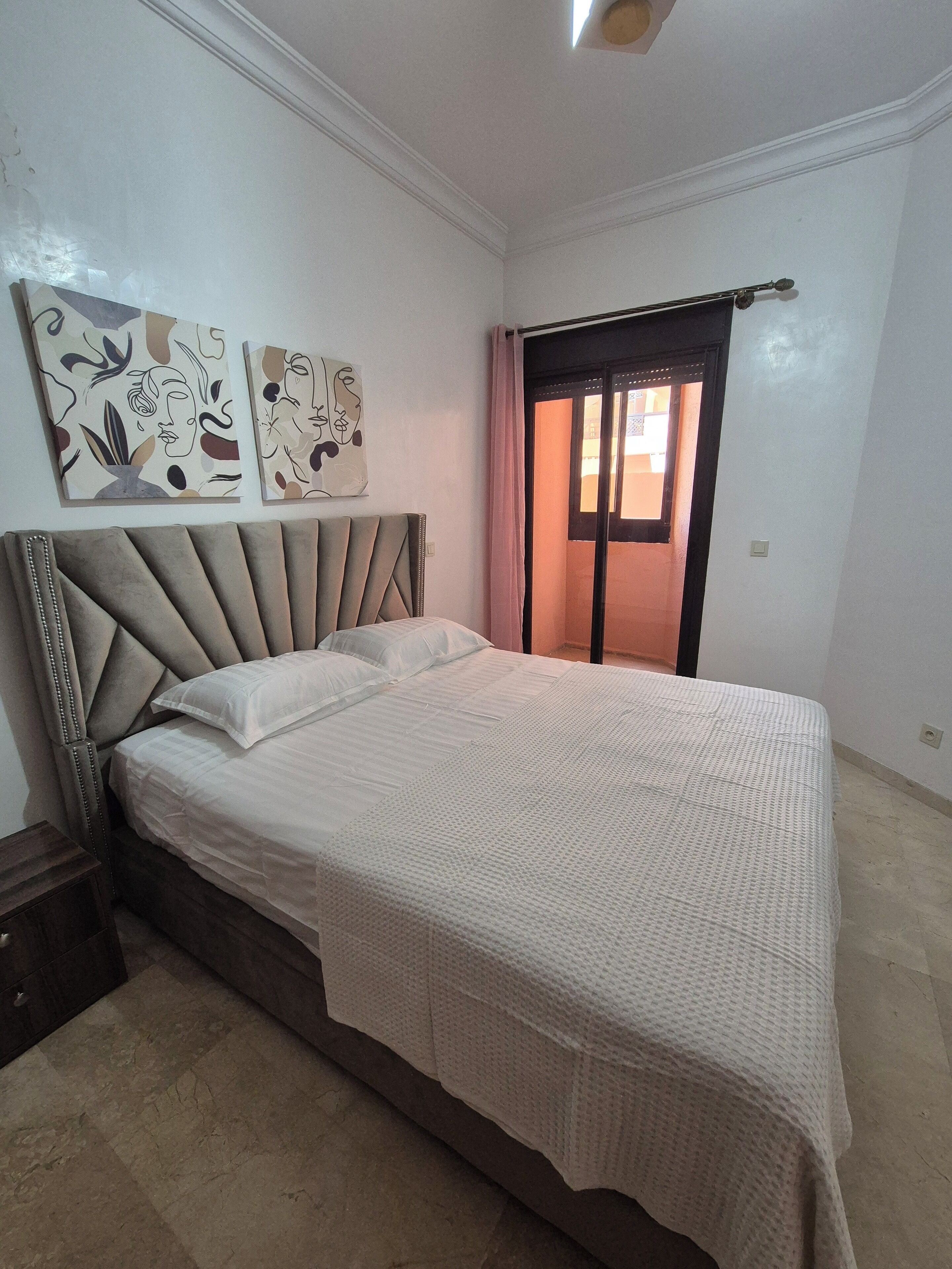 A bedroom with a bed, a nightstand, a window with curtains, and two wall art pieces.