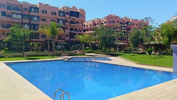Una piscina con superficie de agua cristalina y azul, rodeada de una solana con baldosas. Al fondo, edificios de varios pisos con balcones y ventanas.