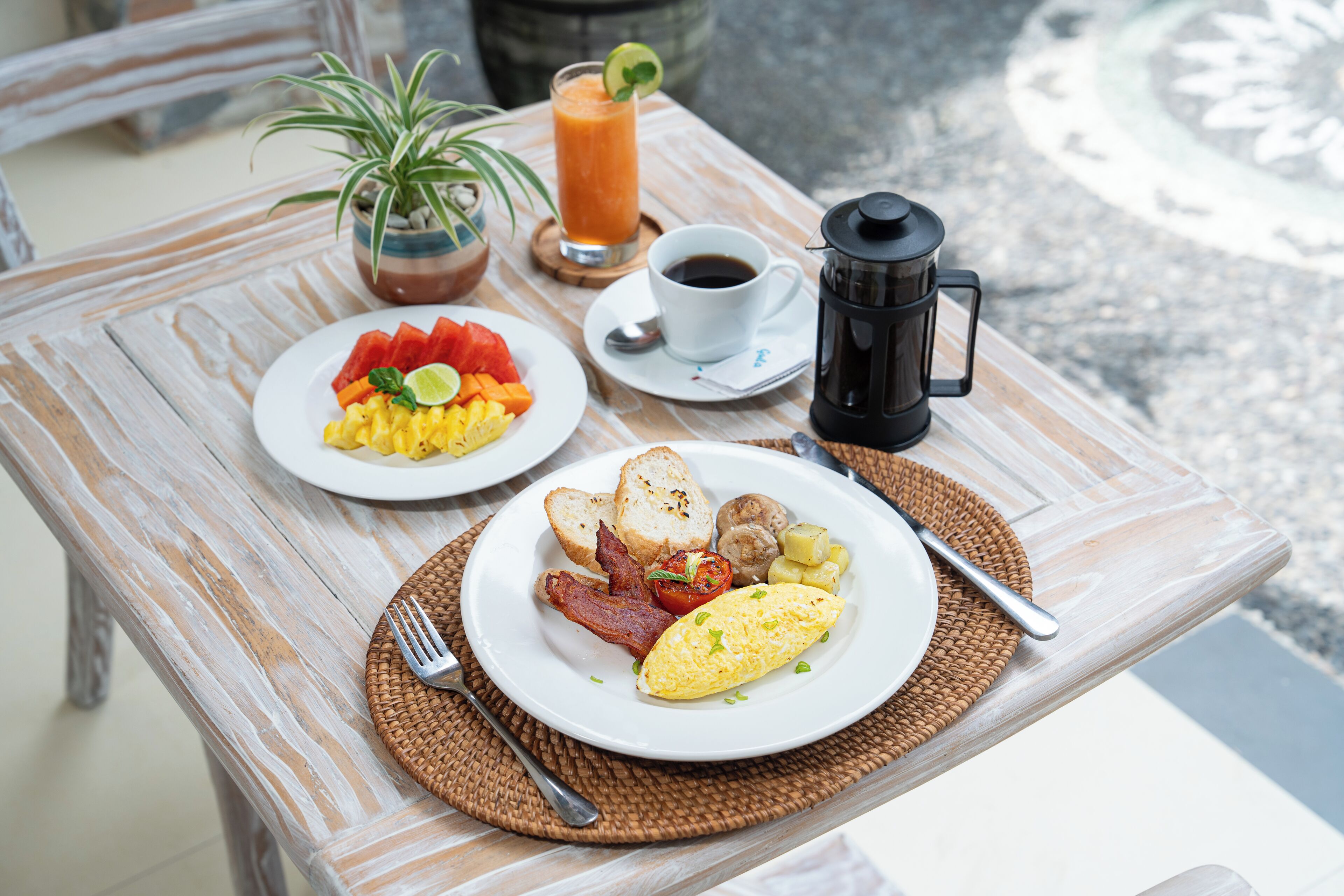 A breakfast table with a plate of eggs, bacon, tomatoes, and toast, a glass of orange juice, and a coffee pot.