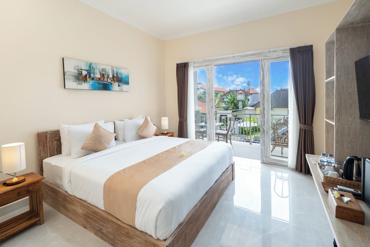 A hotel room with a large bed, a TV, a desk with a chair, and a balcony with a view of buildings and greenery.