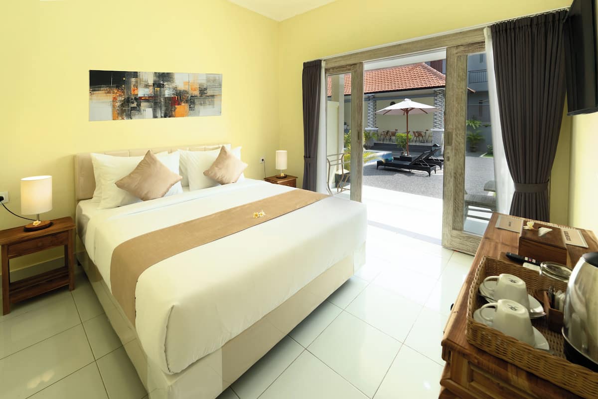 A hotel room with a large bed, a wooden nightstand, a bedside table with a lamp, a small table with a tray of breakfast items, and a view of the outdoor area with a patio and umbrellas.