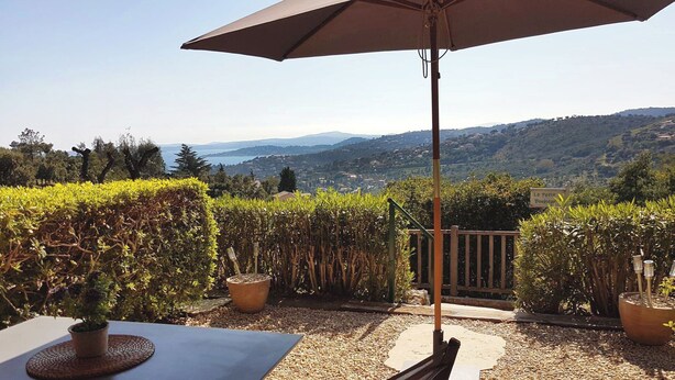 An outdoor patio with a table and chairs, a large umbrella, and a view of a hillside. - Le Petit Bonheur - Studio with sea view - Côte d'Azur (Roquebrune-sur-Argens)