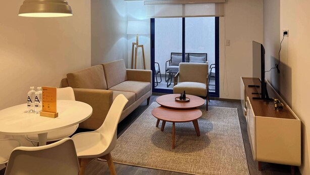 Una sala moderna con un sofá, una mesa redonda y un televisor. Se ve una ventana grande con vistas al exterior.