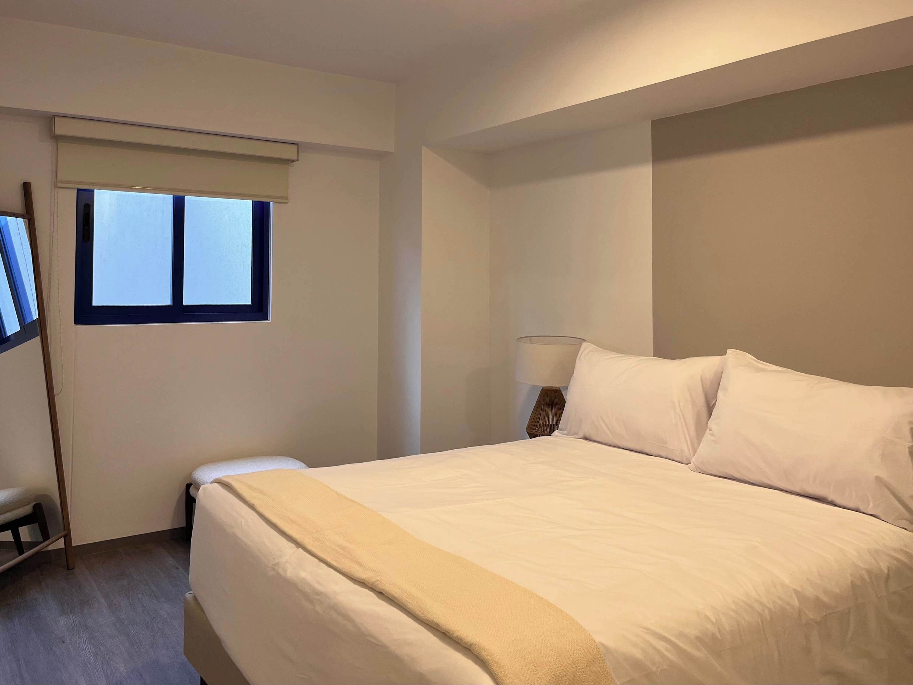 Una habitación de hotel con una cama grande, una mesita de noche con lámpara, una ventana con persianas y una silla.