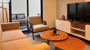 Una habitación de hotel moderna con sofá, sillón, una mesa redonda de madera y televisor sobre un mueble blanco.