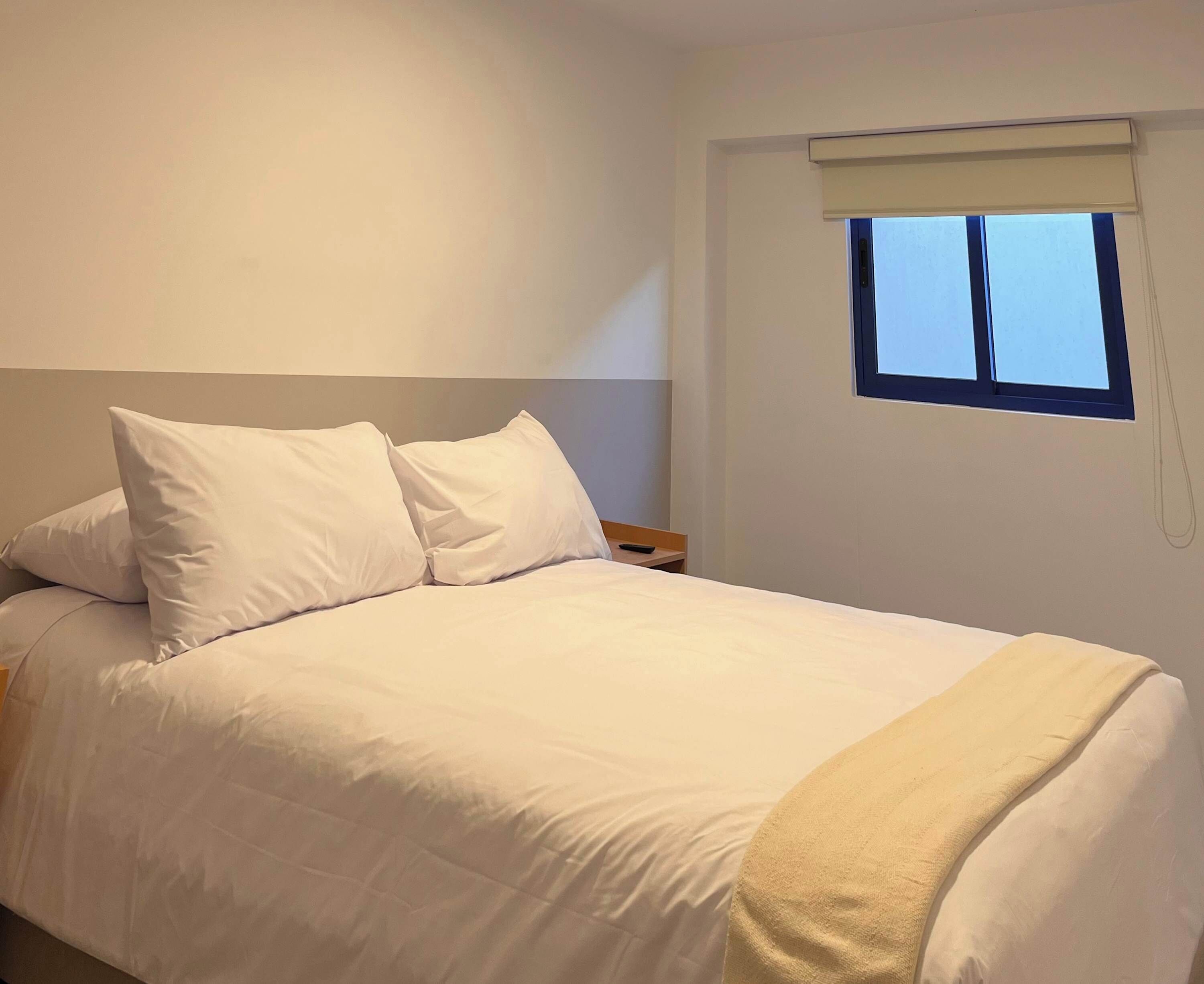 A neatly made bed with white bedding and pillows, a bedside table with a lamp, and a window with a blind.