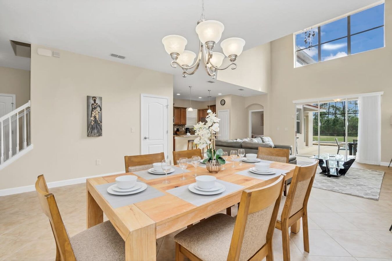 A spacious dining area with a wooden table set for six, surrounded by chairs. A chandelier hangs above, and a painting is visible on the wall.