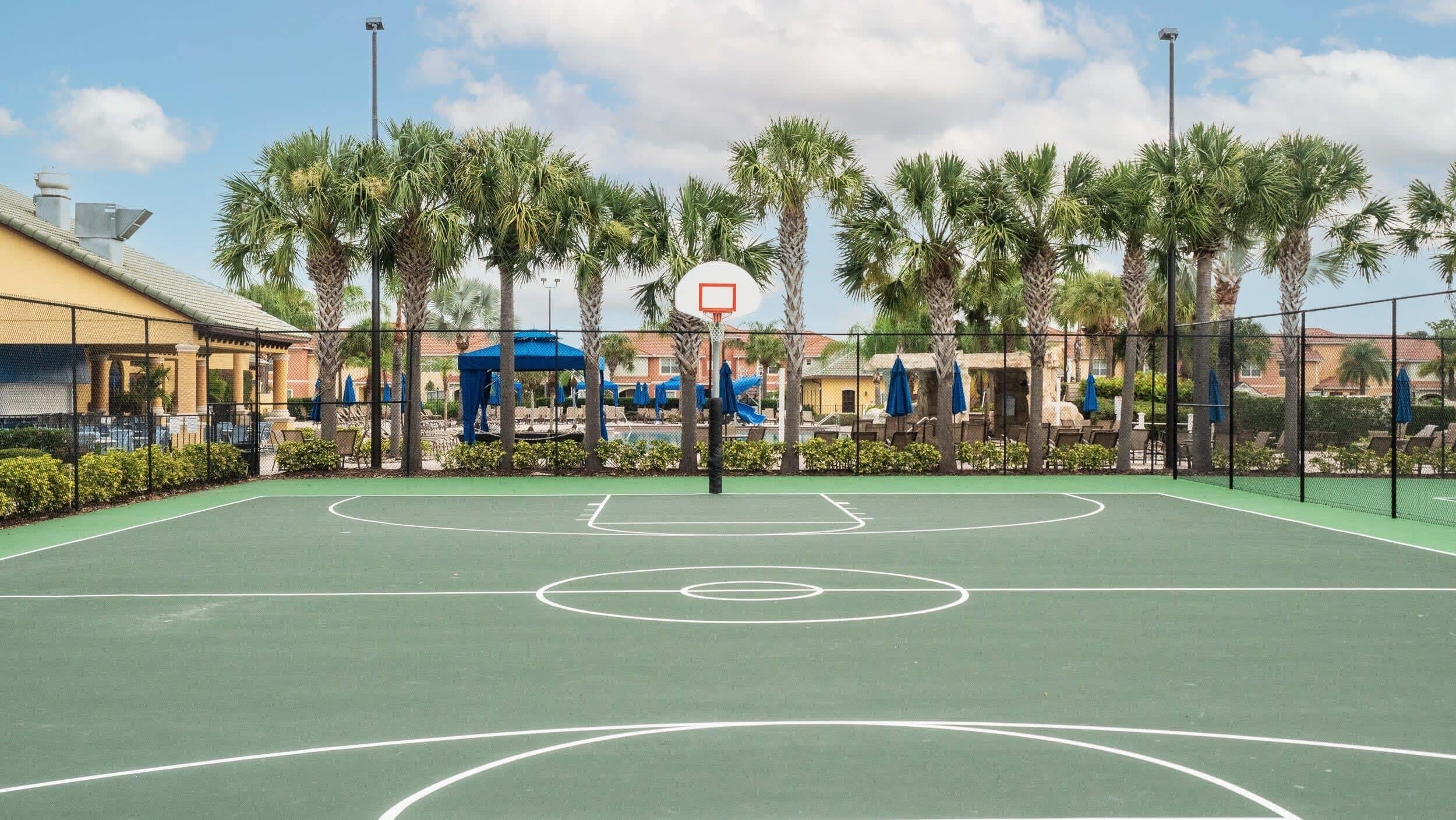 An outdoor basketball court with a basketball hoop, surrounded by palm trees and a fence.