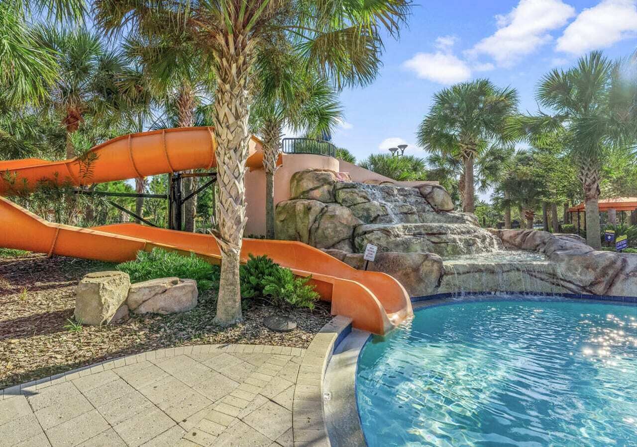Une piscine avec un toboggan aquatique, des palmiers et une cascade artificielle en roche.