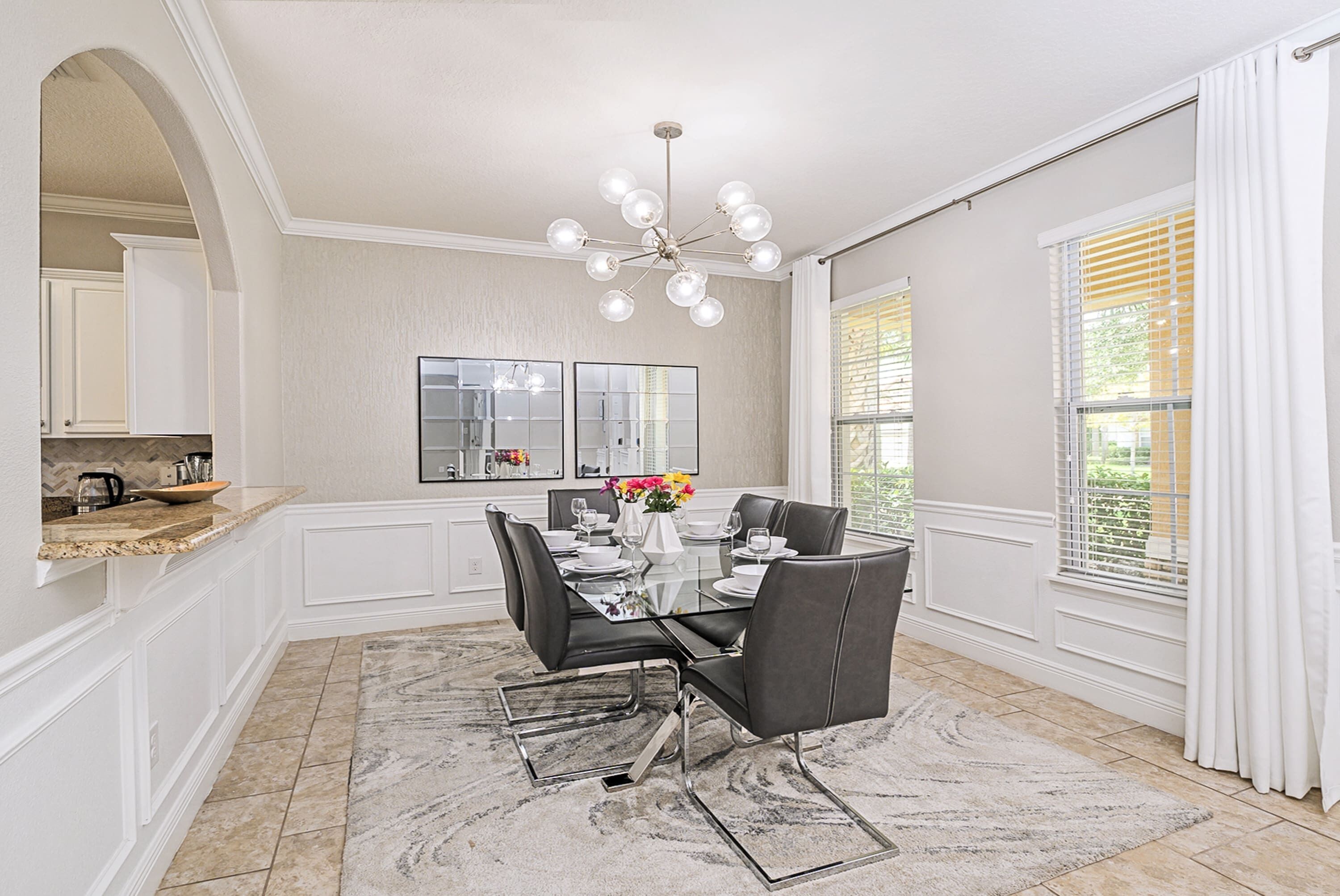 A dining area with a glass table, black chairs, a chandelier, and a view of the outdoors through large windows.