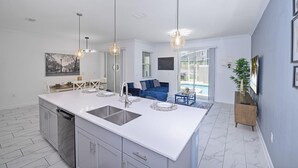 Une cuisine moderne dotée d’un grand îlot central, d’un éclairage suspendu et offrant une vue sur un espace de vie avec un canapé bleu et une piscine.