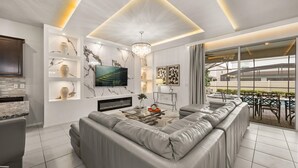 Ein modernes Wohnzimmer mit einer grauen Eckcouch, einem Flachbildfernseher, Marmor-Wandverkleidung und einem großen Fenster mit Blick auf einen Poolbereich.