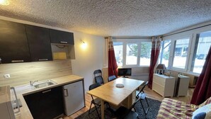 Une cuisine compacte comprenant un évier, un petit réfrigérateur et un coin repas avec une table et des chaises. On y trouve également un téléviseur posé sur un meuble, une fenêtre avec des rideaux et un lit dans un coin.