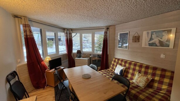 Une pièce chaleureuse avec une table en bois, un canapé et un téléviseur. Il y a des fenêtres avec des rideaux, une lampe et des œuvres d’art accrochées au mur.