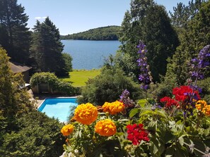 A swimming pool surrounded by lush greenery and vibrant flowers, with a lake and forested hills in the background.