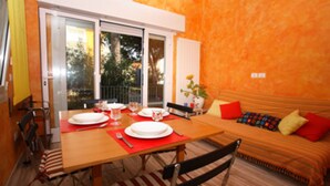 Un coin repas avec une table en bois dressée pour deux personnes, un canapé agrémenté de coussins colorés et une porte-fenêtre coulissante donnant sur un espace extérieur.