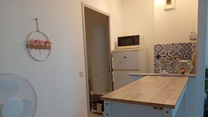A compact kitchen with a microwave on top of a white refrigerator, a small wooden table, and a fan on the left. - Quiet, spacious apartment in an ideal location (Avignon)