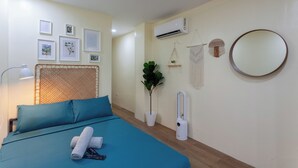 A neatly arranged bedroom with a bed, a bedside lamp, a wall-mounted air conditioner, and framed pictures. - Aesthetic Boho Studio • Near Maginhawa & UP (Quezon City)
