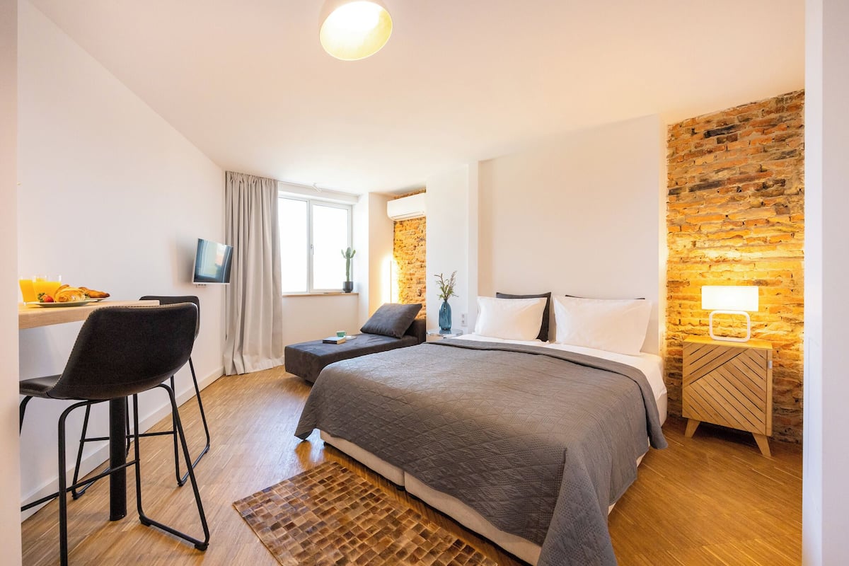 A modern bedroom with a large bed, a desk, and a chair. There is a brick accent wall and a small table with a lamp.
