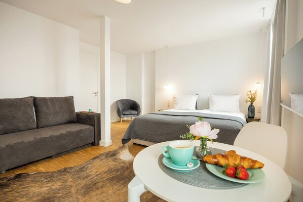 A modern hotel room with a grey sofa, a bed with white linens, a small round table with a cup of coffee and croissant, and a vase with flowers. - Apartments Musala (Varna)
