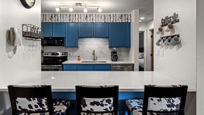 A modern kitchen with blue cabinets, a white countertop, and a dining area with patterned chairs. - Timber Run — Your Perfect Steamboat Getaway, private shuttle to resort (Steamboat Springs)