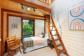 Ein Schlafzimmer mit einem Bett, einem hölzernen Stuhl, einer Leiter, einem Fenster und einer weißen Ziegelwand.
