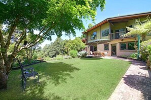 Ein zweistöckiges Haus mit gepflegtem Rasen, einer Terrasse mit Gartenmöbeln und einem großen Baum, der Schatten spendet.