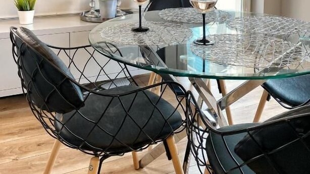 Un coin repas avec une table en verre, des chaises noires, une cheminée où brûle un feu et une fenêtre avec des stores.