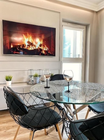 A dining area with a glass table, black chairs, a fireplace with a fire, and a window with blinds.. BP Gray Premium