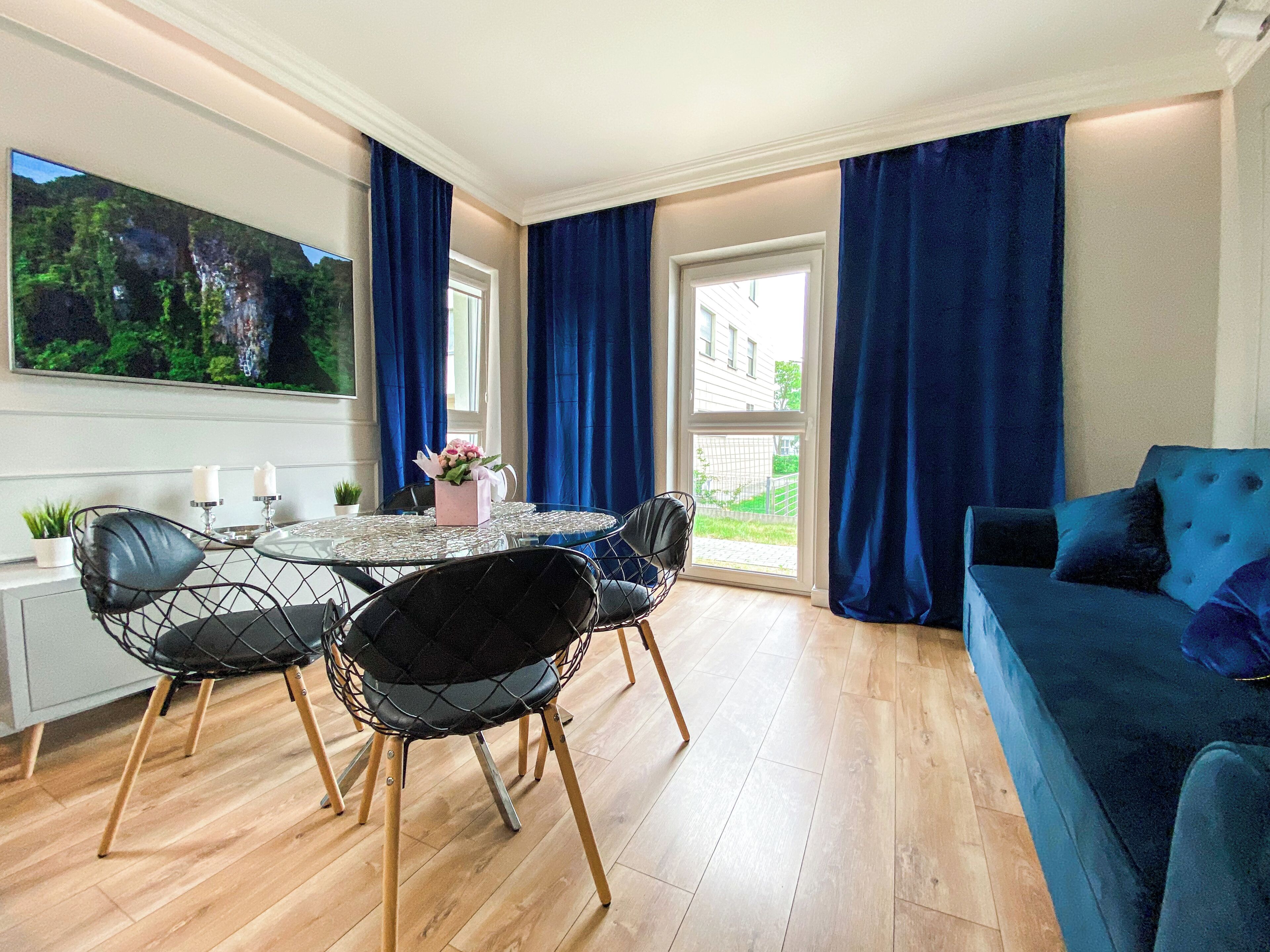 A modern living room with a round glass table, black wireframe chairs, a blue sofa, and a large TV screen.
