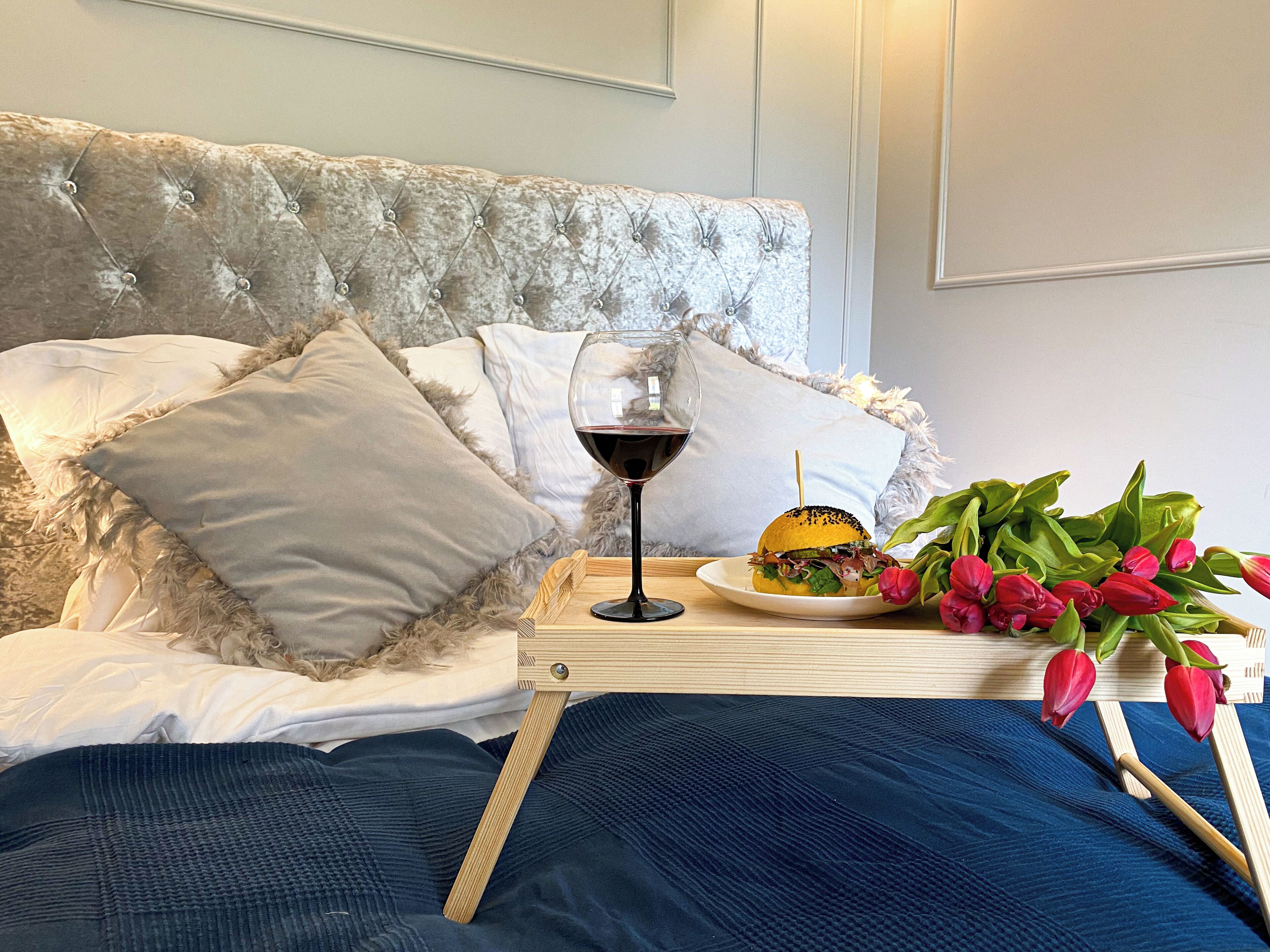 A bed with a tufted headboard, a bedside table with a glass of red wine, a plate with a sandwich, and a bouquet of red tulips.