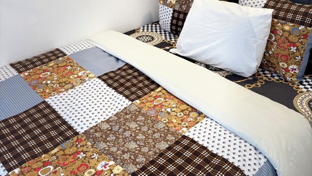 A neatly made bed with a patchwork quilt, a white pillow, and a headboard against a plain wall.